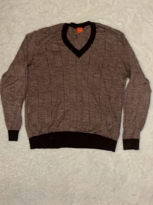 Brun stickad tröja från Hugo Boss - Snygg brun stickad tröja från Hugo Boss med rutigt mönster och V-ringning. Tröjan har svarta ribbade muddar vid ärmslut, nederkant och hals. Perfekt för en stilren och trendig look. Den är äkta och pris kan diskuteras. Den passar även folk som bör storlek M.