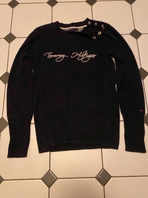 Marinblå stickad tröja Tommy Hilfiger - Snygg marinblå stickad tröja från Tommy Hilfiger med vit broderad logga på bröstet och dekorativa knappar vid axeln. Tröjan har långa ärmar, ribbade muddar och är gjord i ett mjukt stickat material. Perfekt för dig som gillar klassisk och stilren design.