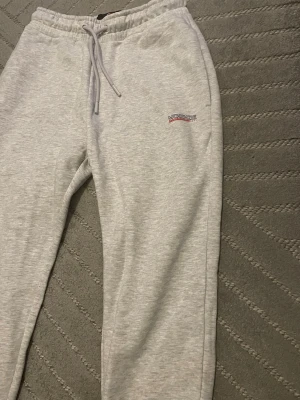 Grå mjukisbyxor från McKenzie - Snygga ljusgrå mjukisbyxor från McKenzie med broderad logga på benet. Byxorna har elastisk midja med snörning och är gjorda i mjukt bomullsmaterial. Perfekta för chill eller träning, med klassisk och enkel stil.