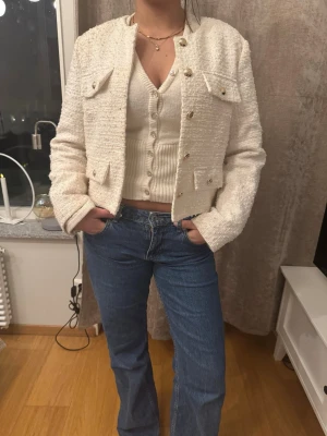 Vit tweed kavaj med knappar - Stilren vit kavaj i tweed-look med dekorativa guldknappar och fyra fickor framtill. Kort, croppad modell som ger en chic vibe och passar perfekt till jeans eller kjol. Klassisk krage och långärmad design för en tidlös look.