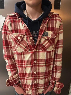 Rödvitrutig skjorta från Superdry - Hejjjj och välkomna till Liljestrands jag säljer nu denna superdry skjortan, han som bär den på bilden är ca 170 men han har hoodie under men även så är den lite stor så skulle rekommendera att du är runt 170 och 180 beror på hur du vill att den sitter kom med frågor och så vidare. så hörs vi. MVH Liljestrand