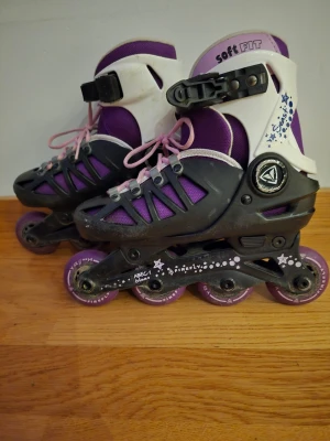 Lila inlines med rosa snören - Säljer ett par snygga inlines från Soft Fit i lila och svart med vita detaljer. De har rosa snören och justerbart spänne vid ankeln. Hjul och stomme är i svart plast med lila hjul. Perfekta för dig som vill cruisa runt i stan eller i parken. Justerbar storlek. 29-32