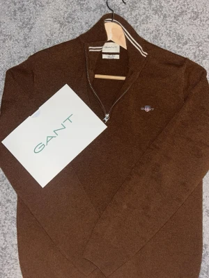 Brun stickad halvzip från GANT - Snygg brun stickad tröja från GANT med halvzip och broderad logga på bröstet. Tröjan har ribbade muddar och en krage med vit rand. Perfekt för lager på lager och ger en clean look till din outfit, väldigt kombinationsfritt enligt mig plus att den är helt oanvänd, luktar oanvänd och har inte en ända fläck överhuvudtaget, köpt för 1599kr på Boozt men säljer för endast 999. Obs materialet är i merinoull.