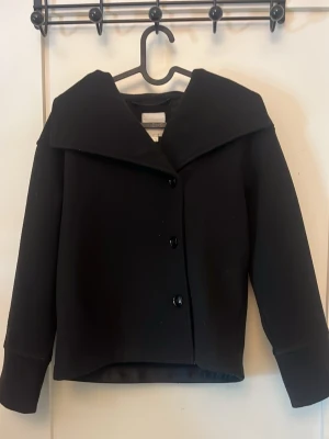 Svart kappa från Gina Tricot - Stilren svart kappa från Gina Tricot med bred krage och tre knappar framtill. Kappan har raka ärmar. Perfekt nu till våren 