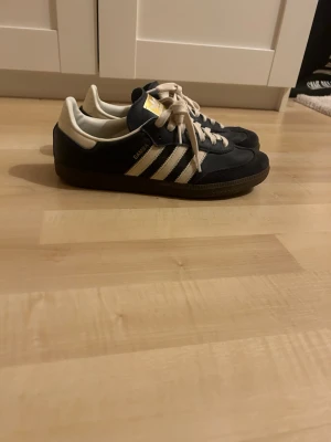 Adidas Samba mörkblå sneakers - Klassiska Adidas Samba sneakers i mrökblå färg med vita läderdetaljer och gummisula. Skorna har de ikoniska tre ränderna på sidan, vit hälkappa och gulddetaljer på plösen. Perfekta för en stilren och sportig look.