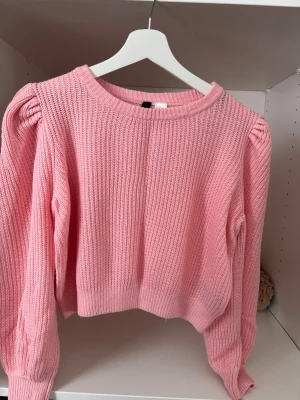 Rosa stickad tröja från Divided - Söt rosa stickad tröja från Divided med puffärmar och ribbstickad struktur. Tröjan har rund halsringning och är lite croppad i modellen. Perfekt för att addera färg till din outfit.