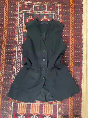 Svart väst med knappar - Klassisk svart väst med v-ringning och fyra knappar framtill. Västen har ett justerbart bälte i ryggen för en snygg siluett. Perfekt att bära över en långärmad tröja💕