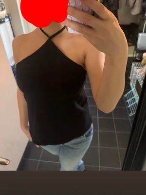 Svart halterneck topp - Säljer en svart halterneck topp med smala axelband som korsas i nacken. Toppens passform är normal och den är ärmlös, perfekt för varma dagar. Aldrig använd!