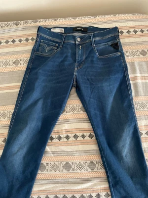 Blå Replay Anbass - Snygga blå jeans från Replay med klassisk femficksmodell och kontrastsömmar. Jeansen har en rak passform och detaljerade bakfickor med broderi samt Replay-lapp i läder bak. Tillverkade i mjukt denimtyg som ger en bekväm känsla.