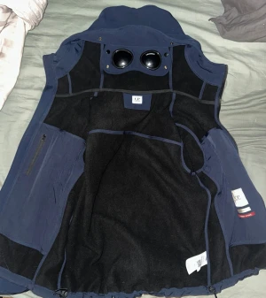 Mörkblå vindjacka med huva från C.P. Company - Säljer en marinblå vindjacka från C.P. Company med ikoniska inbyggda glasögon i huvan. Jackan har dragkedjor framtill, två bröstfickor och justerbar nederkant. Den är i storlek L men jag har M på alla mina andra kläder och den passar fortfarande bra.