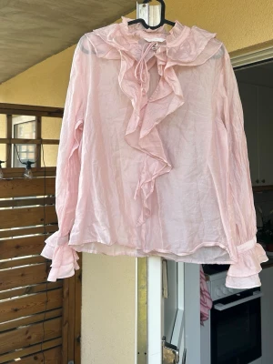 Ljusrosa volangblus från H&M - Supersöt ljusrosa blus från H&M med volangdetaljer längs kragen och framtill. Blusen har långa ärmar med volanger vid ärmslut och knytband vid halsen.🌸endastanvänd en gång, nypris:349