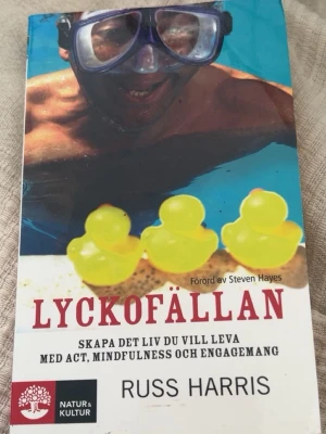 Lyckofällan av Russ Harris - Säljer boken 'Lyckofällan' av Russ Harris, utgiven av Natur & Kultur. Omslaget är färgstarkt med gula badankor och stora röda bokstäver. Boken handlar om ACT-metoden och mindfulness, och är en lättillgänglig självhjälpsbok.