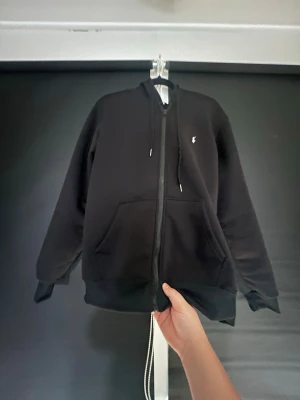 Svart zip hoodie med liten logga - Snygg svart hoodie med dragkedja och huva. Har en liten vit logga på bröstet och stora fickor framtill. Klassisk passform och ribbade muddar vid ärmar och nederkant. Perfekt för en chill och avslappnad stil.