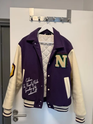 Lila college jacka Zara - Snygg bomberjacka från Zara i lila och vitt med broderade detaljer och stora bokstäver på bröstet. Jackan har quiltat foder, ribbade muddar och klassisk college-stil. Perfekt för dig som vill sticka ut med en cool och sportig look.