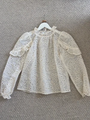 Vit broderad blus med volangdetaljer från neo noir - Säljer en vit blus med broderade hålmönster och volangdetaljer på axlarna. Blusen har långärmade puffärmar. Blusen är från neo noir och köpt för 749kr. Ny utan prislapp då den inte kommit till användning sen jag köpte den.