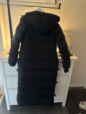 Svart lång dunjacka från Canada Goose - Säljer en svart lång dunjacka från Canada Goose med huva och klassisk logga på ärmen. Jackan är quiltad och har dragkedja framtill. Perfekt för kalla vinterdagar och håller dig varm med sin rejäla dunfyllning.