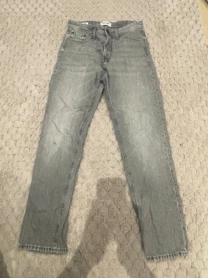 Grå raka jeans i klassisk modell - Säljer ett par grå jeans med rak passform och fem fickor. Jeansen har en klassisk look med ljus tvätt och normal midja. Perfekta för en avslappnad stil.
