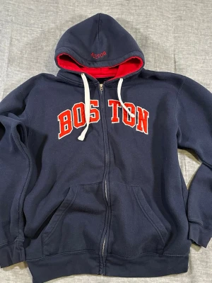 Boston spell out hoodie zip navy red collegiate L - Bay State Gear Boston hoodie with classic varsity spell out. Navy fleece with red hood lining. Full zip, kangaroo pockets, relaxed fit. Nice everyday collegiate look. Two letters have small stitch repair (shown in pics) but overall solid.  Size L P2P 57 cm Length 70 cm Sleeve 62 cm S2S 62 cm Waist 50 cm