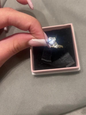 Guldfärgad ring med stora stenar - Säljer en guldfärgad ring med två stora, gnistrande stenar i olika former framtill. Ringen har även små stenar längs bandet för extra bling. Perfekt för dig som gillar statement-smycken och vill sticka ut lite extra.