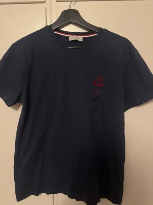Mörkblå Moncler T-shirt - Snygg mörkblå t-shirt från Moncler med fyra loggor i rött och blått tryckta vertikalt på både fram- och baksidan. Klassisk rund halsringning och korta ärmar. Tillverkad i mjuk bomull för skön känsla och stilren look.