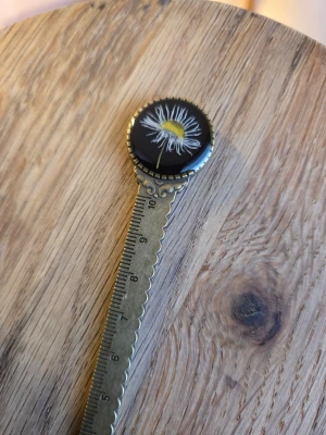 Blommig bokmärke/linjal i metall - Unikt bokmärke i metall med en rund topp som har en vit och gul blomma mot svart bakgrund. Bokmärket har dekorativa kanter och ingraverade siffror längs skaftet. Perfekt för dig som vill ha något snyggt och annorlunda till böckerna.