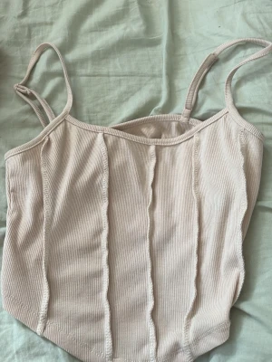 Beige ribbad linnetopp med sömmar - Trendig beige linnetopp med ribbad struktur och dekorativa synliga sömmar framtill. Tunn, stretchig och med smala axelband. Perfekt att styla med jeans eller kjol för en enkel men snygg look.