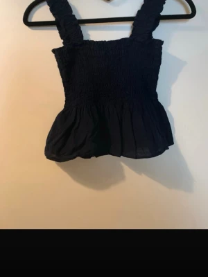 Svart smockad linnetopp med volang - Supersöt svart linnetopp med breda smockade axelband och volang nertill. Perfekt för dig som gillar en feminin och trendig stil. Toppens smockade material ger en bekväm och snygg passform. Har en liten fläck.