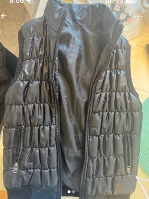 Svart quiltad dunväst med dragkedja - Snygg svart dunväst med quiltat mönster och hög krage. Västen har två framfickor med dragkedja och ribbade detaljer vid ärmöppningarna och nederkanten. Perfekt att slänga över en hoodie för extra värme. Materialet är glansigt och ger en modern look.