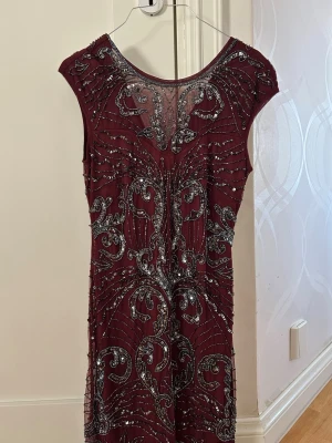 Vinröd glittrig kortklänning - Vinröd kortklänning med glittriga silverdetaljer och vackert mönster. Klänningen har ärmlös design, rund halsringning fram och v-ringning bak. Perfekt för bal eller festliga tillfällen. Materialet känns lyxigt och skimrande.