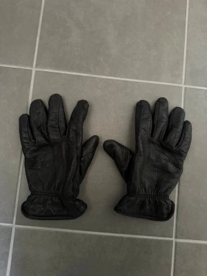 Svarta skinnhandskar från Hestra, stl 9 - Svarta handskar i mjukt skinn med resår vid handleden för bättre passform. Klassisk design med fem fingrar, perfekta för kyliga dagar. Snygga och stilrena, passar till de flesta outfits.