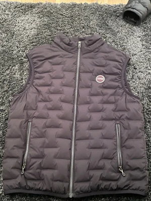 Svart dunväst från Colmar - Snygg svart dunväst från Colmar med quiltat mönster och hög krage. Västen har två dragkedjeförsedda fickor framtill och en logga på bröstet. Perfekt att slänga över en hoodie för en chill look. Materialet är lätt och värmande.