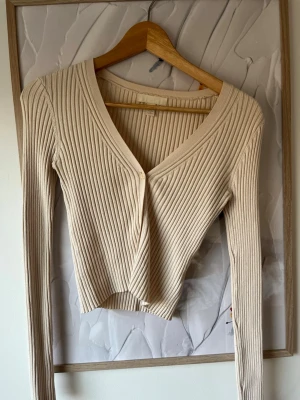 Beige ribbad omlottkofta - Trendig beige ribbad omlottkofta med djup V-ringning och lång ärm. Kortare modell med en knapp framtill. 