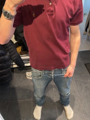 Vinröd pikétröja från Polo Ralph Lauren - Snygg vinröd pikétröja från Polo Ralph Lauren med klassisk krage och knappar framtill. Tröjan har korta ärmar och den ikoniska broderade loggan på bröstet. Perfekt för en stilren och avslappnad look.