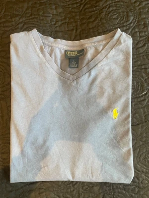 Grå v-ringad t-shirt från Polo Ralph Lauren - Snygg grå t-shirt från Polo Ralph Lauren med v-ringning och klassisk gul broderad logga på bröstet. Tillverkad i mjuk bomull som känns skön mot huden. Perfekt för en clean och stilren look. Postas dagen eller dagen efter köpet.