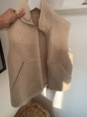 Beige teddyväst med dragkedja - Mysig beige teddyväst med hög krage och dragkedja framtill. Västen har två snedställda fickor och är gjord i ett fluffigt, fårskinnsinspirerat material som ger en trendig look. Perfekt att slänga över en hoodie eller tröja för extra värme.