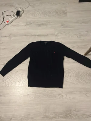 Mörkblå kabelstickad tröja Polo Ralph Lauren - Snygg mörkblå kabelstickad tröja från Polo Ralph Lauren med rund halsringning och diskret röd logga broderad på bröstet. Tröjan har långa ärmar och ribbade muddar vid ärmslut och nederkant. Perfekt för lager på lager och klassisk stil.