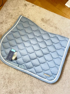 Ljusblått schabrak från Lami-Cell - Aldrig använt! Säljer ett ljusblått schabrak från Lami-Cell med quiltat mönster och vit kant. Ordinarie pris 700kr. Tillverkat i glansigt tyg som ger en snygg look till din hästutrustning. Perfekt för dig som vill ha stil på ridningen. Märke i metall på ena sidan.