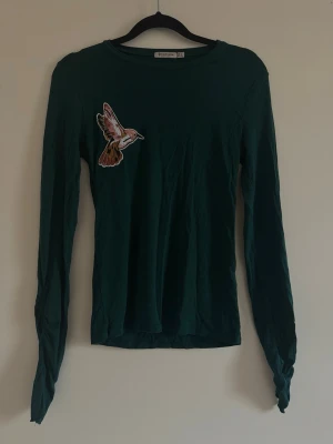 Mörkgrön långärmad topp med fågel - Mörkgrön långärmad topp med en snygg broderad fågelpatch på bröstet. Toppen har rund halsringning och är i mjuk bomull, perfekt för en chill och trendig look.