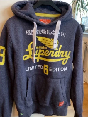 Mörkblå Superdry hoodie med tryck(L fits M) - Snygg mörkblå hoodie från Superdry med stor gul logga och japansk text på bröstet. Har vita dragsnören i huvan, känguruficka och gult nummertryck på ena ärmen. Mjuk insida och ribbade muddar. Perfekt för en avslappnad streetwear-look. har en liten fläck på armen