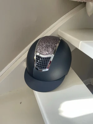 Crw Marinblå ridhjälm med glitterdetalj - Ordinarie pris 1 500kr. Snygg Marinblå ridhjälm från Crw med glittrig panel framtill och silvriga detaljer. Hjälmen har en klassisk skärm. Perfekt för dig som vill sticka ut i stallet och samtidigt ha bra skydd. Strl M 56-58, använda ca 10 gånger, aldrig tappade eller trillat av med. Har två stycket likadana, kolla sista bilden, har också en till annons på den andra. Pris kan absolut diskuteras, skriv om du har några frågor!