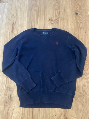 Mörkblå sweatshirt från Ralph Lauren L (14-16) - Klassisk mörkblå sweatshirt från Ralph Lauren med rund halsringning och den ikoniska röda logotypen broderad på bröstet. Tröjan har ribbade muddar vid ärmslut och nederkant, perfekt för en avslappnad och stilren look.