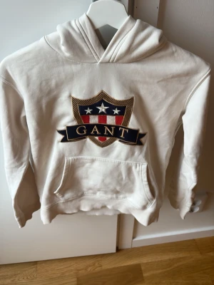 Vit hoodie med GANT-logga - Vit hoodie från GANT med stor broderad sköldlogga och text på bröstet. Klassisk känguruficka framtill och huva. Mjuk bomullskänsla och ribbade muddar vid ärmslut och nederkant. Perfekt för en avslappnad och stilren look.