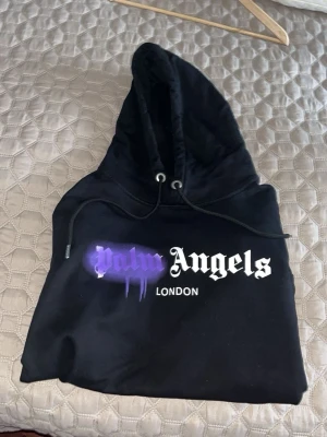 Palm Angels svart hoodie London - Snygg svart hoodie från Palm Angels med stor huva och dragsko. Framsidan har ett tryck med texten 'Palm Angels London' där 'Palm' är i lila graffiti-stil. Perfekt för dig som gillar streetwear och vill sticka ut med en unik design.
