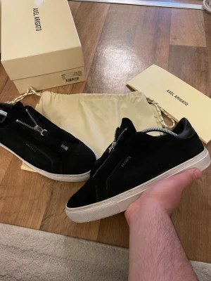 Svarta sneakers från Axel Arigato - Stilrena svarta sneakers från Axel Arigato i mocka med vit sula och dragkedja istället för klassiska snören. Diskret logga på sidan och minimalistisk design. Perfekta för dig som gillar en clean look och vill sticka ut lite extra. Storlek 41 passar 42