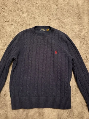 Mörkblå kabelstickad tröja Polo Ralph Lauren - Kabelstickad mörkblå tröja från Polo Ralph Lauren med rund halsringning och röd broderad logga på bröstet. Tröjan har långa ärmar och ribbade muddar vid ärmslut och nederkant. Perfekt för lager på lager och klassisk stil.
