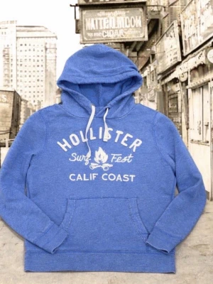 Blå hoodie från Hollister - Blå hoodie från Hollister med vit text och tryck på bröstet där det står 'Surf Fest Calif Coast'. Hoodien har huva med vita snören och en stor magficka framtill. Tillverkad i mjukt material, perfekt för en avslappnad stil. Står som XL men passar L