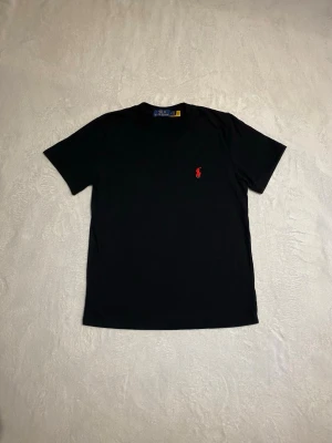 Svart t-shirt från Polo Ralph Lauren - Klassisk svart t-shirt från Polo Ralph Lauren med röd broderad logga på bröstet. T-shirten har rund halsringning och korta ärmar. Tillverkad i mjuk bomull som känns skön mot huden. Perfekt basplagg för en clean och stilren look.