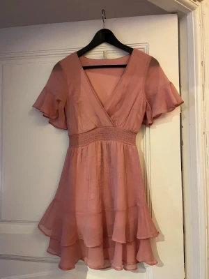 Rosa omlottklänning med volanger - Söt kort rosa klänning med omlott-skärning och v-ringning. Klänningen har korta volangärmar, smock i midjan och dubbla volanglager i kjolen för extra flow. 