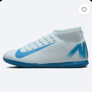 Nike vita och blå fotbollsskor - Snygga fotbollsskor från Nike i vitt med blå detaljer och logga. Skorna har en högre krage för extra stöd och är tillverkade i syntetmaterial med mönstrad yta för bättre bollkontroll. Perfekta för spel på inomhusplaner.