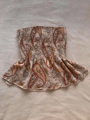 Beige paisleymönstrad linnetopp - Supersöt linnetopp i beige med paisleymönster i rött, gult och brunt. Smockad upptill för en tight passform över bysten och volang nedtill som ger en flowy känsla. Tunna axelband med spetsdetalj.